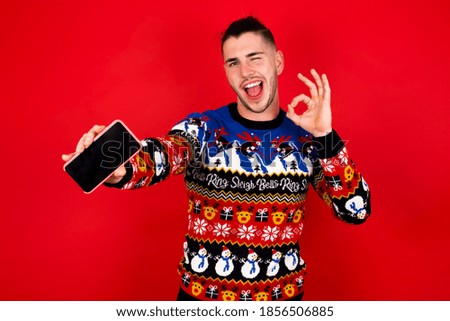 Young handsome Caucasian man wearing Christmas sweater against red wall holding in hands cell showing ok-sign