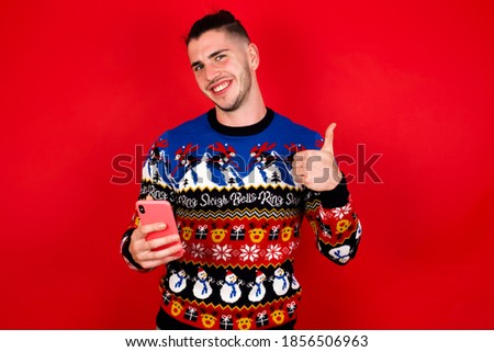 Portrait of Young handsome Caucasian man wearing Christmas sweater against red wall using and texting with smartphone  happy with big smile doing ok sign, thumb up with fingers, excellent sign