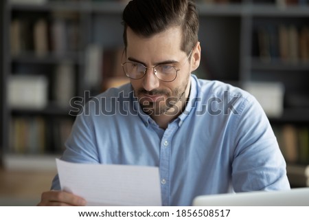 Serious focused man sitting at workplace desk holding paper sheet reading news in correspondence learn written information in formal business letter, legal notification from bank, got proposal concept Royalty-Free Stock Photo #1856508517