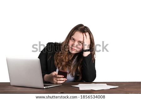 Cute office girl posing while working at her work table. Laptop and some papers placed in front of her. She dressed in strict black suit. She feels tired and a bit puzzled. Isolated, white background.