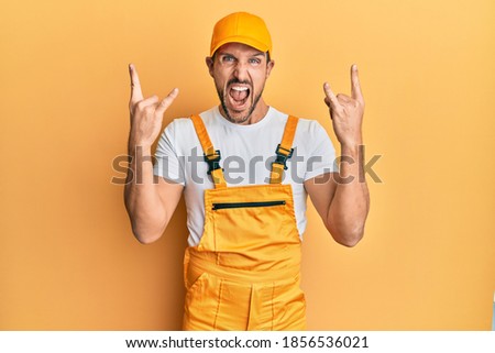 Young handsome man wearing handyman uniform over yellow background shouting with crazy expression doing rock symbol with hands up. music star. heavy music concept. 
