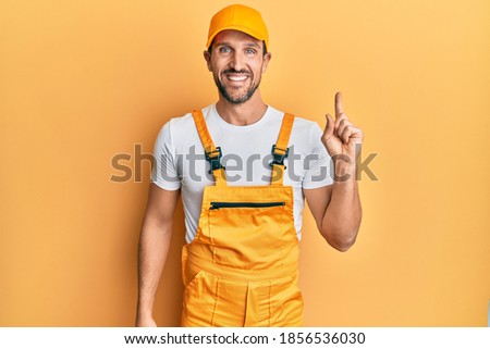 Young handsome man wearing handyman uniform over yellow background with a big smile on face, pointing with hand finger to the side looking at the camera. 
