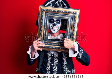Young man wearing mexican day of the dead costume holding empty frame pointing finger to one self smiling happy and proud 