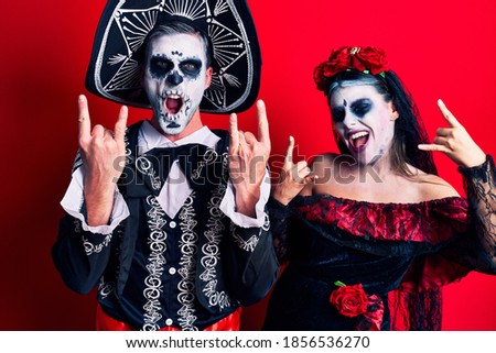 Young couple wearing mexican day of the dead costume over red shouting with crazy expression doing rock symbol with hands up. music star. heavy concept. 
