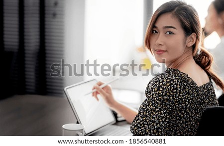 Portrait of young beautiful asian woman working on tablet computer, smiling and looking at camera while sitting at the table in modern office.