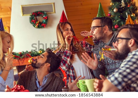 Group of friends sitting by nicely decorated Christmas tree, having fun while celebrating Christmas and friend's birthday at home, blowing party whistles