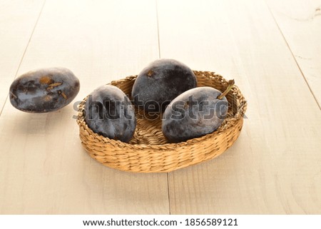 Ripe dark purple plums in a straw plate, close-up, on a wooden table.