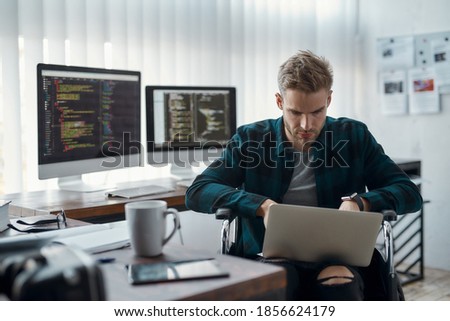 Young focused male web developer in a wheelchair writing program code on his laptop at his workplace with multiple computer screens on the background. Disability concept. IT software