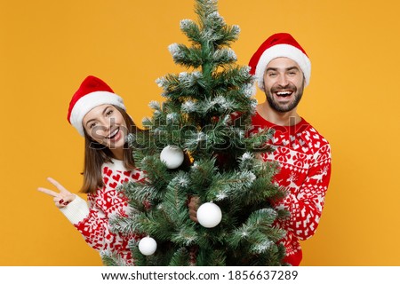 Funny cheerful young Santa couple friends man woman in sweater Christmas hat hold fir tree showing victory sign isolated on yellow background studio. Happy New Year celebration merry holiday concept