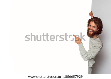 Young handsome  man with ginger beard hiding behind the banner. Looking at camera. Perfect happy smile.   Copyspace.