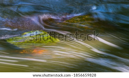 Nature background of a wild stream detail flowing over mossy stones. Abstract green and blue dynamic wavy water surface with motion blur on white streamlines. Close-up of fast running swift creek bed.