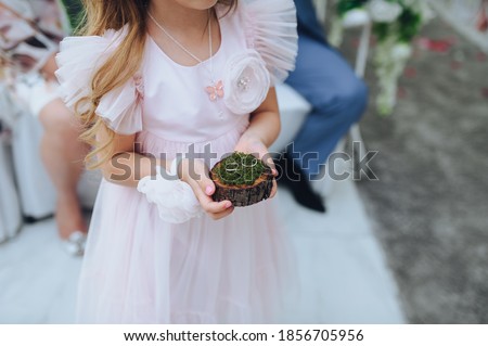 A little girl in a pink dress carries wedding rings on a wooden moss stand. A touching moment at the ceremony. Hands and rings.  Royalty-Free Stock Photo #1856705956