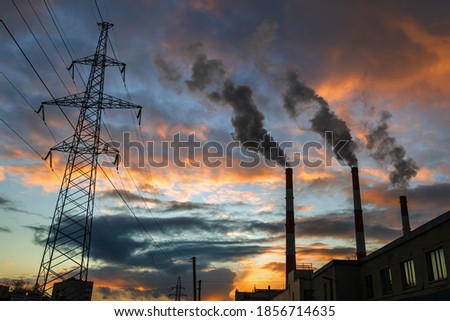 Silhouette of steel lattice tower, three chimneys smoke on dramatic sunset sky background. Transmission tower and industry pipes. Industrial cityscape