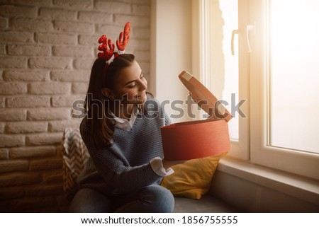 A cheerful young teenage girl with a deer-ears prop on her head peeks into a red gift box with a ribbon as she sits on the couch by the window. Celebrating the New Year and Xmas at home