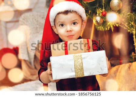 Cute little boy in a santa hat and with christmas gifts in his hands sits under the christmas tree
