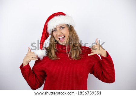 Young pretty blonde woman wearing a red casual sweater and a christmas hat over white background shouting with crazy expression doing rock symbol with hands up