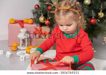 toddler girl with headband with horns of a deer sitting near the Christmas tree, opening a gift.