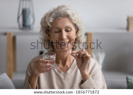 Head shot smiling mature woman holding glass of water and white round pill in hands, sitting on couch at home, overjoyed satisfied middle aged female taking supplements, healthcare concept Royalty-Free Stock Photo #1856771872
