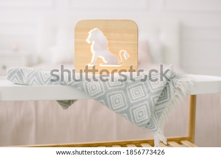 Front close up view of stylish wooden night lamp with lion cut out picture, on gray blanket at cozy light bedroom interior. Wooden decorations at home interior
