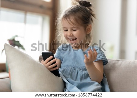 Cheery little girl watching cartoons on smart phone. Kid girl sit on couch hold gadget spend time on internet fun website for children. New generation and modern tech overuse, parental control concept Royalty-Free Stock Photo #1856774422