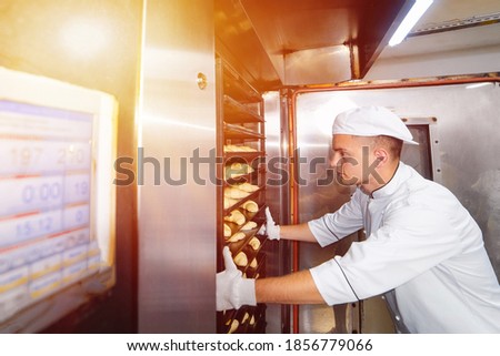 baker boy inserts a cart with raw dough baking trays into an industrial oven in a bakery. Royalty-Free Stock Photo #1856779066