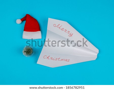 White paper airplane with the inscription: merry christmas, santa hat and christmas tree on a blue background, top view close-up.