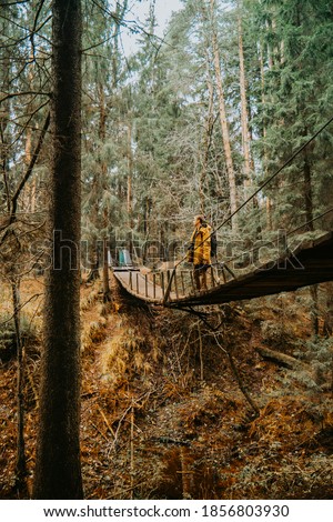 A young photographer in a yellow jacket sits on a suspension bridge and takes pictures of the river beneath him. A man in the middle of a suspension bridge.