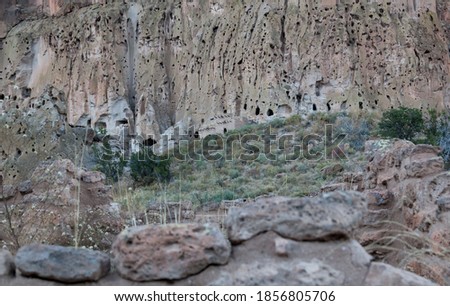 A building made of brick and clay with wooden poles to support a roof matches the sandstone background of the cliff walls of Frijoles Canyon in Bandelier National Monument, New Mexico.