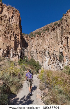 A man with a camera, tripod, and backpack walks on a pathway in Frijoles Canyon looking at the rock walls with cave dwellings in Bandelier National Monument, New Mexico.