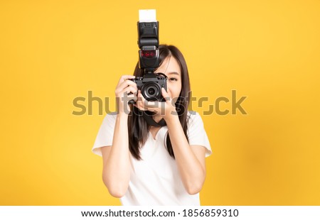 Happy smiling asian young girl photographer and looking viewfinder on retro digital mirrorless photo camera ready to shoot at studio shot on yellow background.