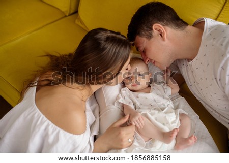 Young caucasian beautiful parents mom and dad hug and kiss their newborn daughter sitting on the sofa at home
