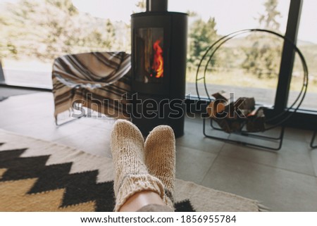 Feet in warm knitted woolen socks on background of modern black fireplace and big windows with view on mountains. Woman relaxing in comfortable home, cozy warm moments