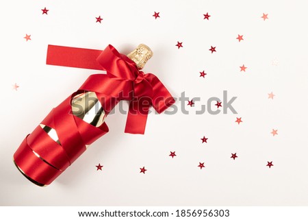 a gift bottle of champagne in golden foil wrapped in a red silk ribbon and scattered confetti on a white background