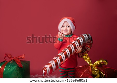 Toddler boy in Santa hat with Xmas gifts and candy cane on a red background.