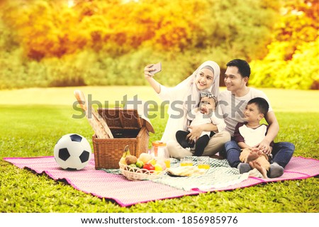 Muslim family using a cellphone to taking a selfie photo together while picnicking in the park with autumn trees background