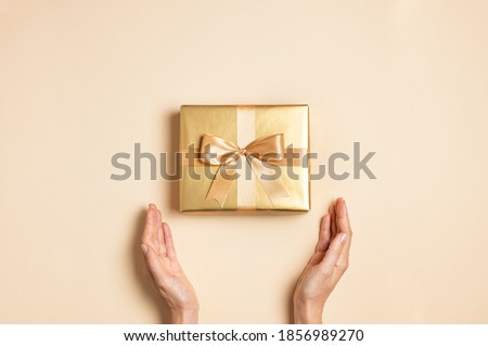 Female hands hold Golden gift with ribbon bow on beige background flat lay top view copy space. Holiday present, christmas, new year, birthday, valentine's day, 8 march. Surprise Royalty-Free Stock Photo #1856989270