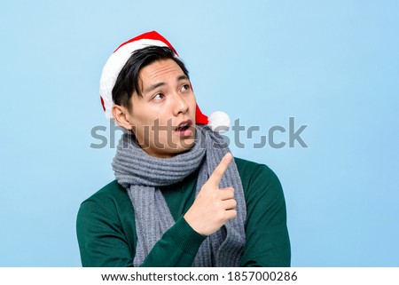 Surprised Asian man in Christmas attire looking and pointing hand up to empty space aside on light blue isolated background