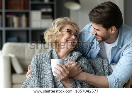 Smiling mature woman holding cup, hugging with adult son close up, enjoying leisure time, happy elderly mother wearing warm scarf drinking hot tea or coffee, family spending weekend together at home Royalty-Free Stock Photo #1857042577