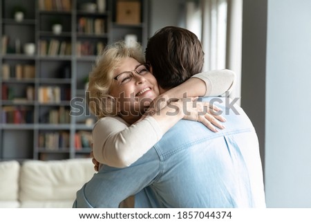 Close up smiling mature woman wearing glasses hugging adult son, standing in living room at home, family enjoying tender moment, beautiful happy middle aged woman embracing young man Royalty-Free Stock Photo #1857044374