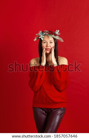 full-length portrait of a smiling Asian young woman with a Christmas wreath, red lipstick and hands on her face on a scarlet background.