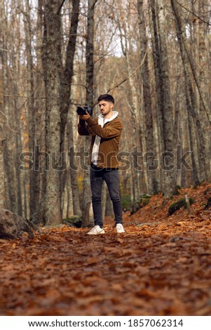 Caucasian boy in beautiful forest with red leaves taking a picture with professional reflex camera photography student with brown jacket, white t-shirt and red scarf, it's cold