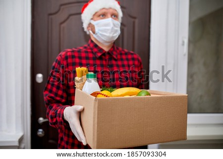 Volunteer man in Santa hat and protective mask and gloves delivery donation box at home in Christmas