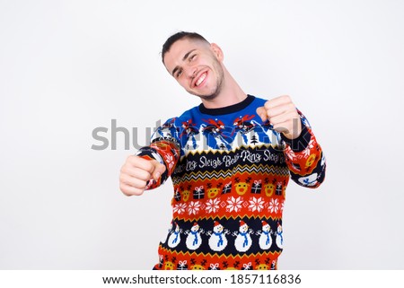 Portrait of charming Young handsome Caucasian man wearing Christmas sweater against white wall, smiling broadly while holding hands over her head.  Confidence and relax concept.