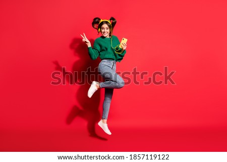 Full size photo of young cheerful positive girl show v-sign listen music in headphones hold mobile isolated on red color background