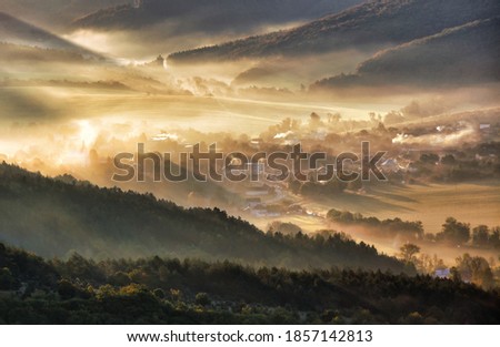 Aerial view of village in fog with golden sunbeams at sunrise in autumn. Beautiful rural landscape with road, buildings, foggy colorful trees. Slovakia
