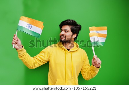 Urban young hipster indian man in a fashionable yellow sweatshirt hold flags of India in hands. Cool south asian guy wear hoodie isolated on green background.