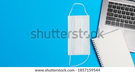 Flat lay composition with laptop, medical face mask, notepad on blue background top view with copy space. Concept of safety work in coronavirus pandemic.