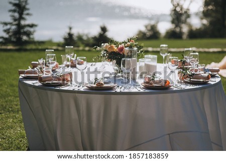 A large round table with a white tablecloth and dishes, a bouquet of flowers stands on the green grass in the park in nature. Wedding ceremony, party. Holiday decorations.