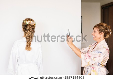 Beautiful blonde woman dressed in white on her wedding day before getting married, her mother takes pictures of her with smart phone.