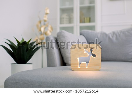 Wooden hand made accessories. Wooden night lamp with deer picture, on gray cozy soft sofa, at stylish light home living room interior. Home decor and lamps.